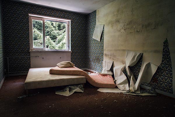 A broken down apartment bedroom with the wallpaper peeling off and the mattress coming off the frame