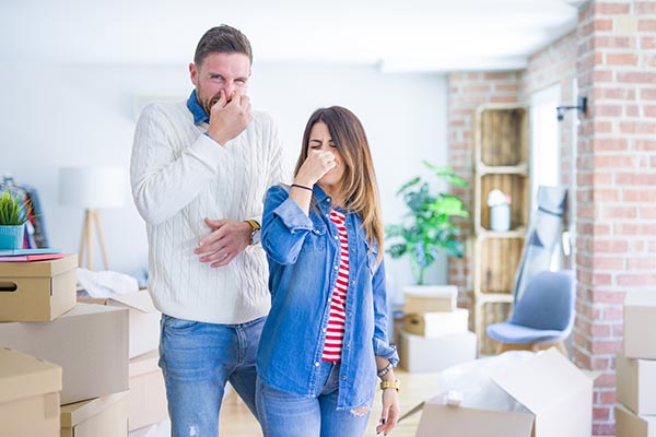 Young beautiful couple standing at new home around cardboard boxes smelling something stinky and disgusting, intolerable smell, holding breath with fingers on nose. Bad smells concept.