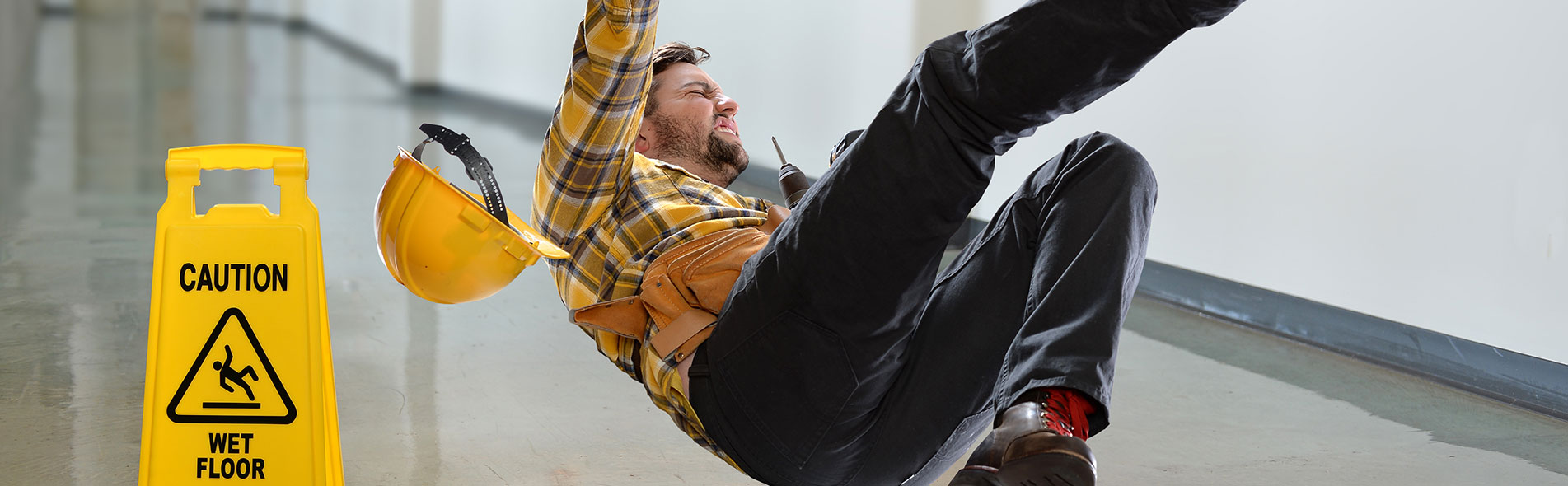 A man in a hallway slips and falls, his yellow helmet flying off his head