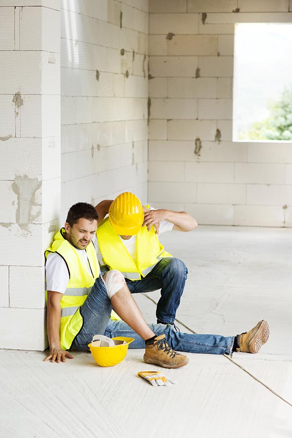 A man winces in pain while sitting against the wall, getting help from a co-worker after suffering a work injury